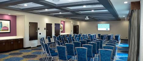 Ein Konferenzraum mit Reihen blauer Stühle, einer Projektionsleinwand und einem gemusterten Teppich.