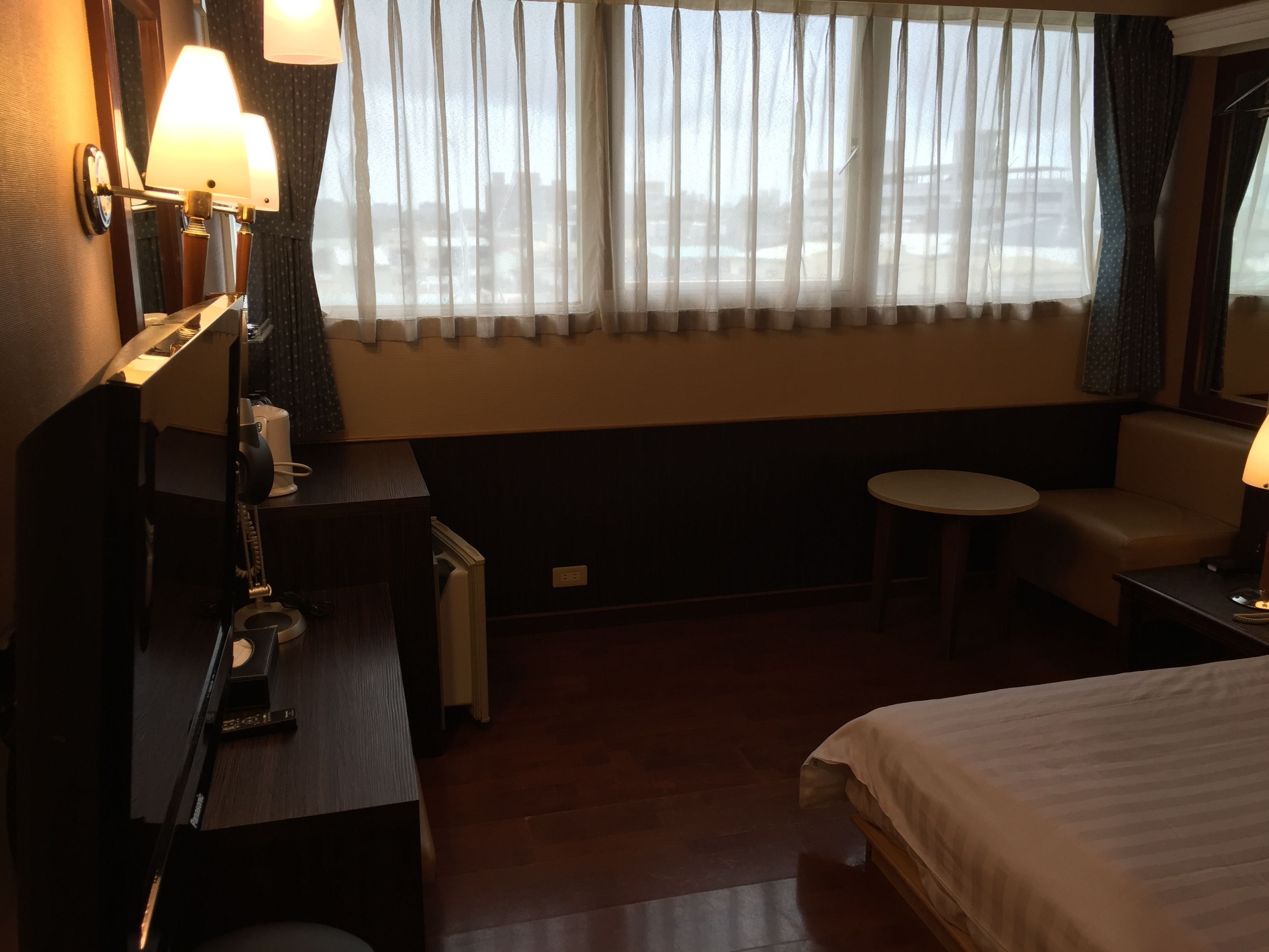 Una camera d'albergo con un letto, una scrivania, una sedia, un tavolino e una finestra con tende leggere.