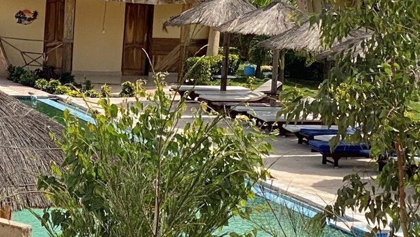 Un área de piscina con sombrillas de paja, sillas de descanso y un edificio con puertas.