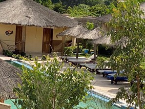A pool area with thatched umbrellas, lounge chairs, and a building with doors. - Hotel & Campement du Lac Rose (Niaga)