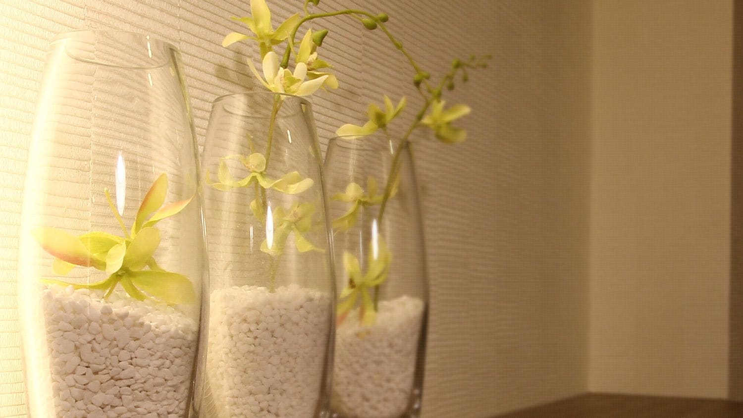 Tre glasvaser med vita småsten och gula blommor, placerade framför en vägg med en tydlig struktur.