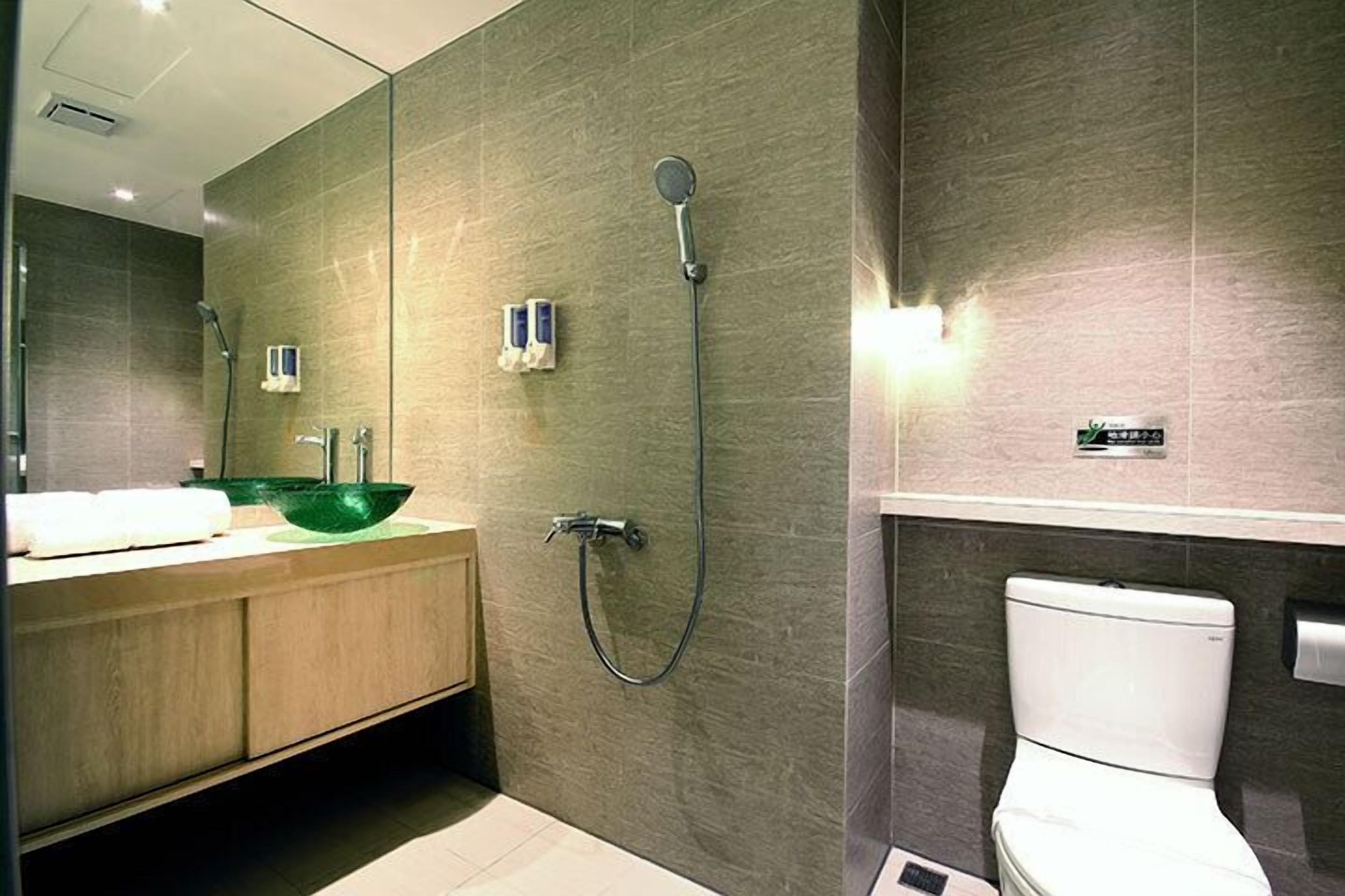 A modern bathroom with a large mirror, a green bowl sink, a shower, and a toilet.