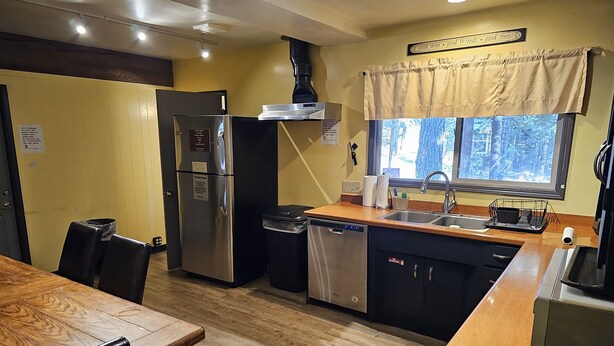 A compact kitchen with a refrigerator, dishwasher, and a window with a view of trees. - Moderne Hostel (Mammoth Lakes)