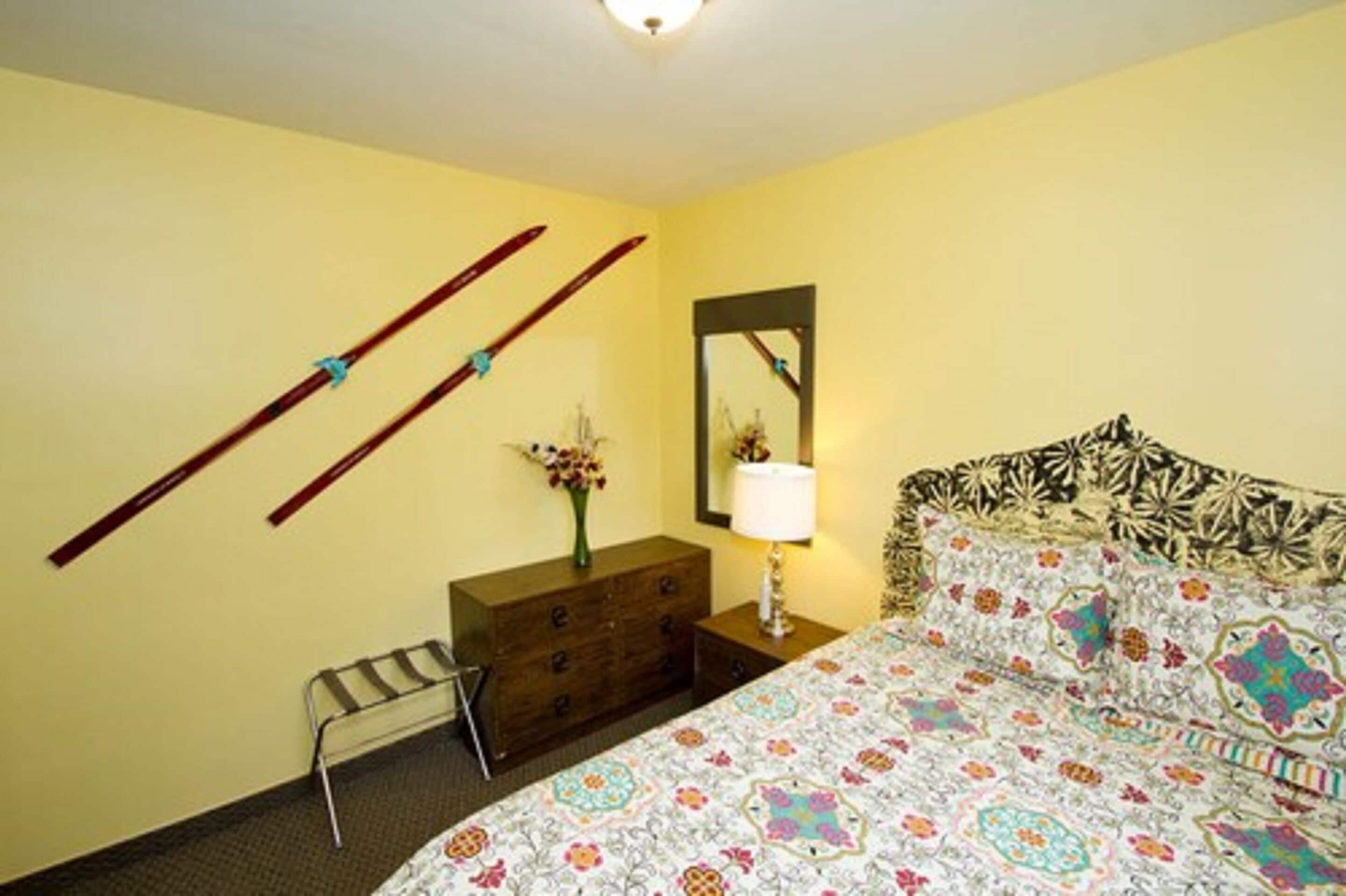 Une chambre à coucher avec un lit, une commode, un miroir et des skis appuyés contre le mur.