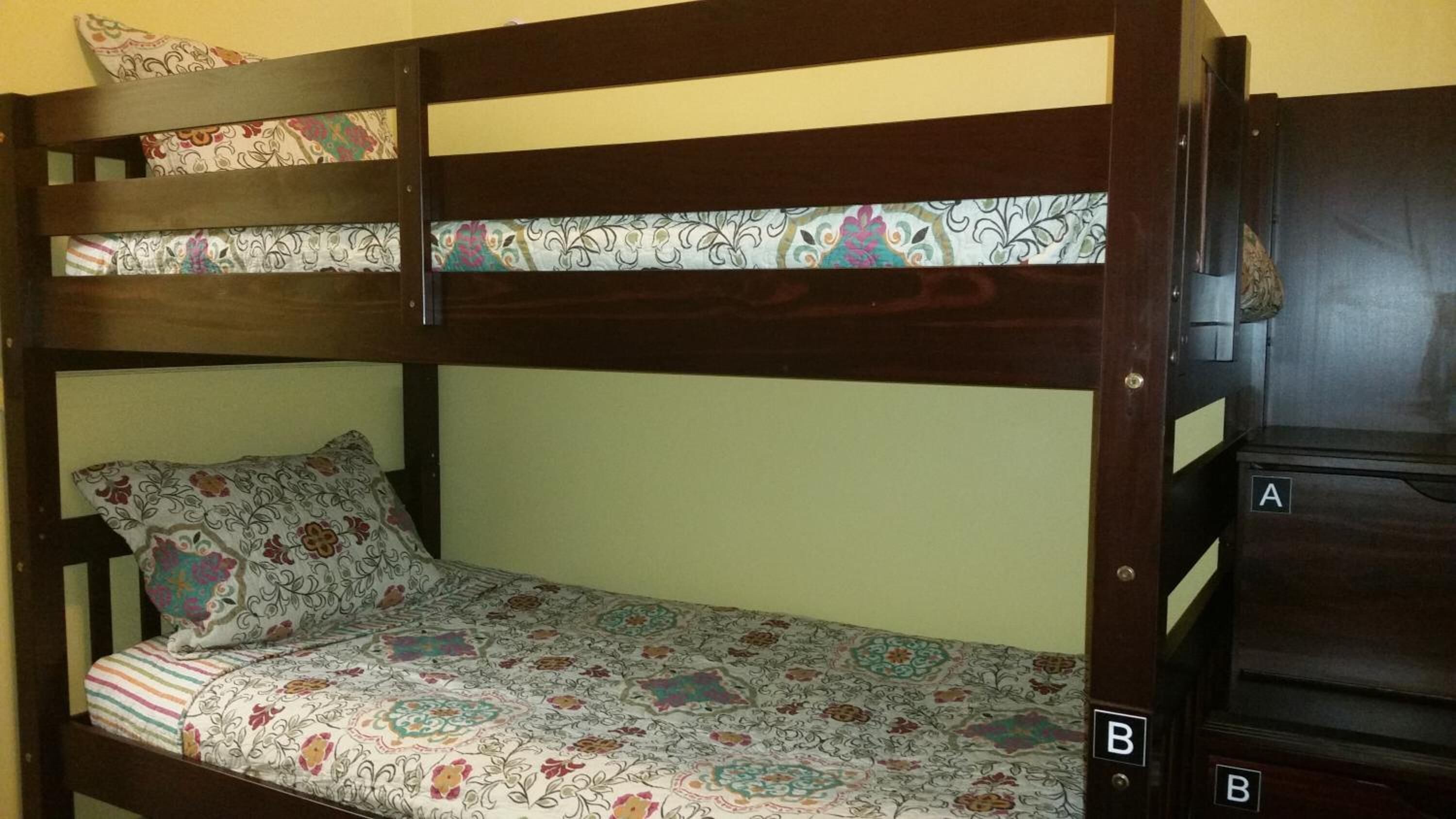 Una cama litera con colcha y almohadas estampadas, cabecera de madera y un pequeño cofre con las etiquetas 'A' y 'B'.