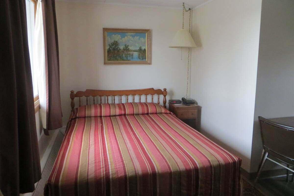 A room with a bed, a bedside table with a lamp, a chair, and a painting on the wall.