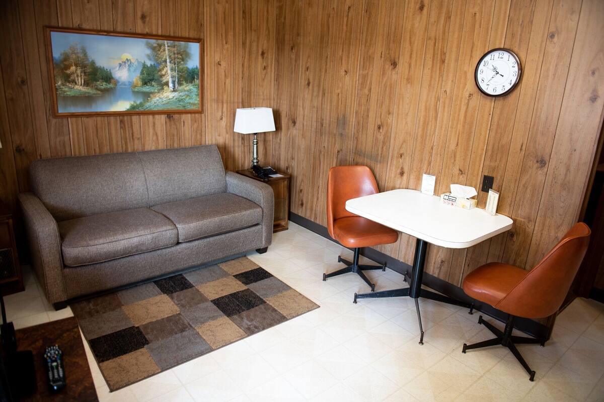 A room with a sofa, a small table with chairs, a clock, and a painting on the wall.