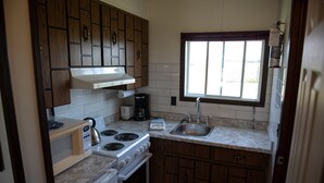 A compact kitchen with wooden cabinets, a built-in oven, a stovetop, and a window providing natural light. - Adams Golden Acres Motel (Kingsville)