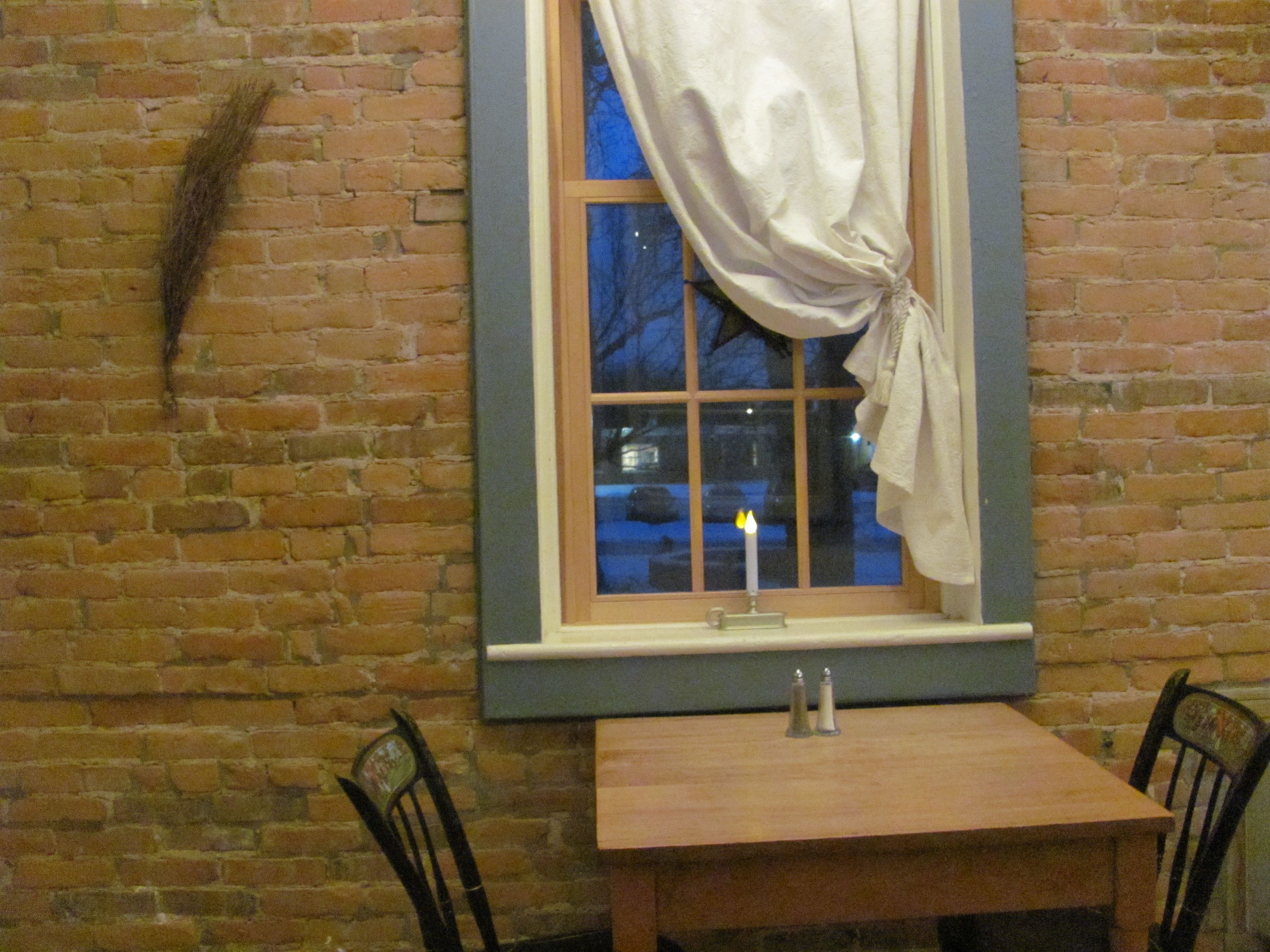 A brick wall with a window featuring a white curtain tied back, a wooden table, and two black chairs.