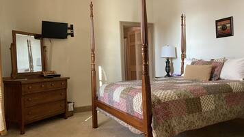 A bedroom with a four-poster bed, a dresser with drawers, a mirror, a television mounted on the wall, and a door leading to another room.