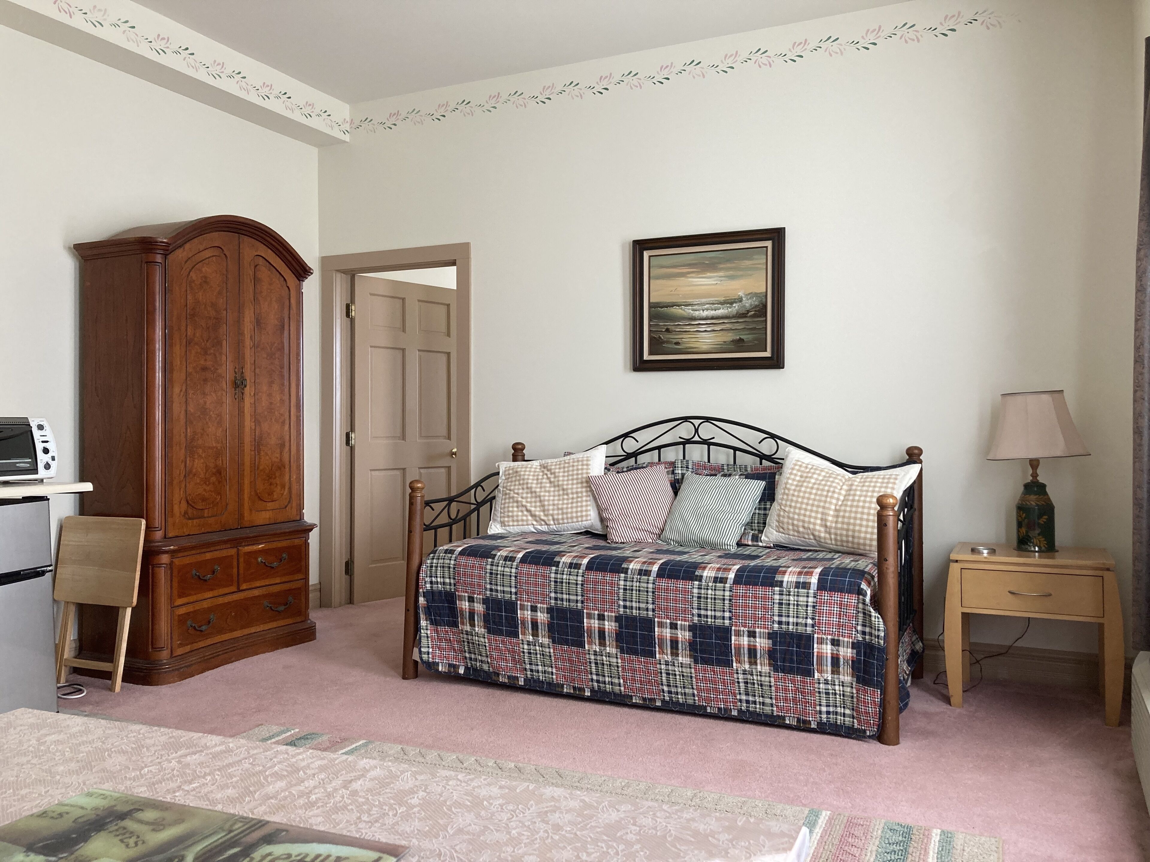 A bedroom with a bed, a wooden wardrobe, a desk with a computer, and a bedside table with a lamp.
