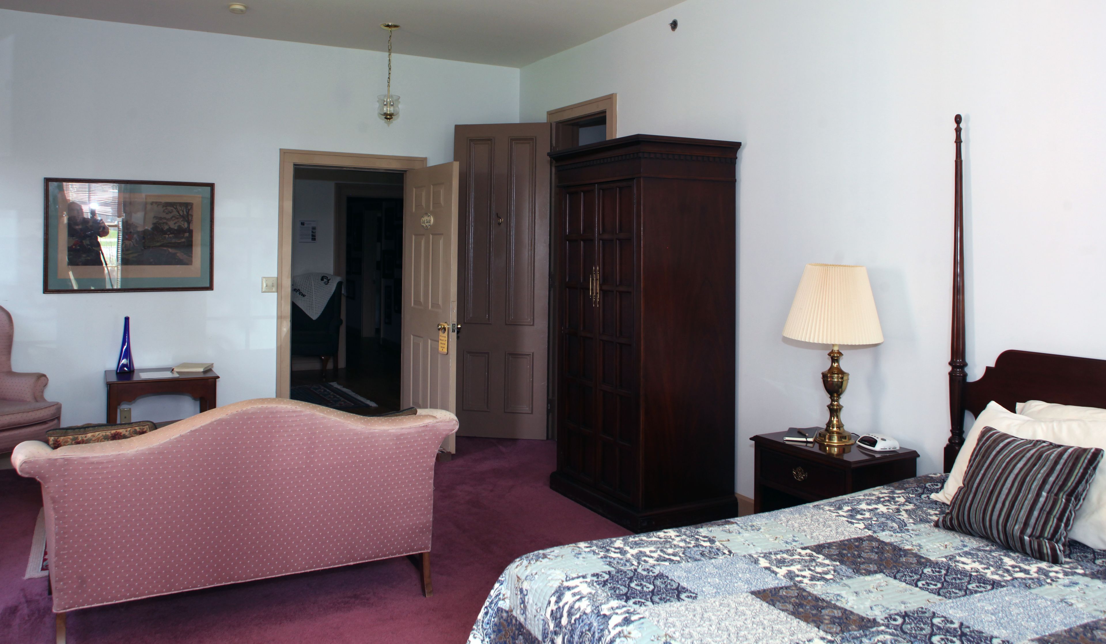 Un dormitorio con una cama, un tocador, una silla, una lámpara y una puerta que da a otra habitación.