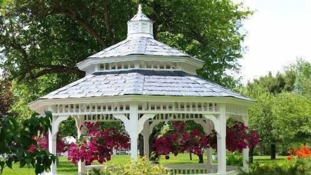 Een witte pergola met een puntige kap, omringd door weelderige begroeiing en bloeiende planten.