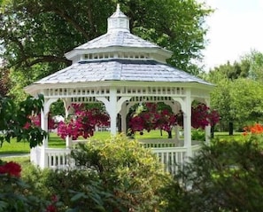 Un belvédère blanc, doté d’un toit en pente, est entouré d’une végétation luxuriante et de plantes en fleurs.