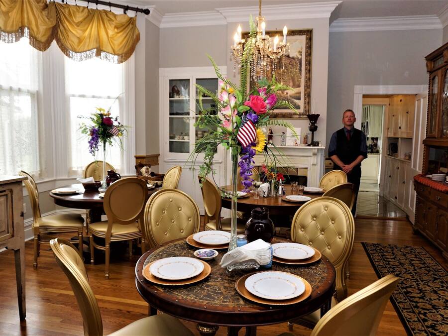 Comedor con una mesa redonda dispuesta para cuatro personas, rodeada de sillas tapizadas. Un candelabro cuelga encima y se ve un ventanal con cortinas.