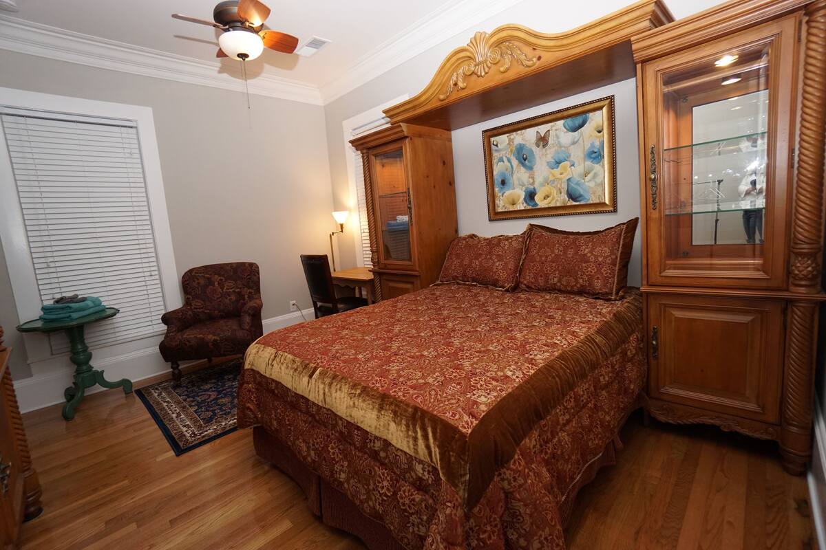 Une chambre à coucher avec un grand lit, une tête de lit en bois, un ventilateur de plafond, un tableau et une armoire en bois.