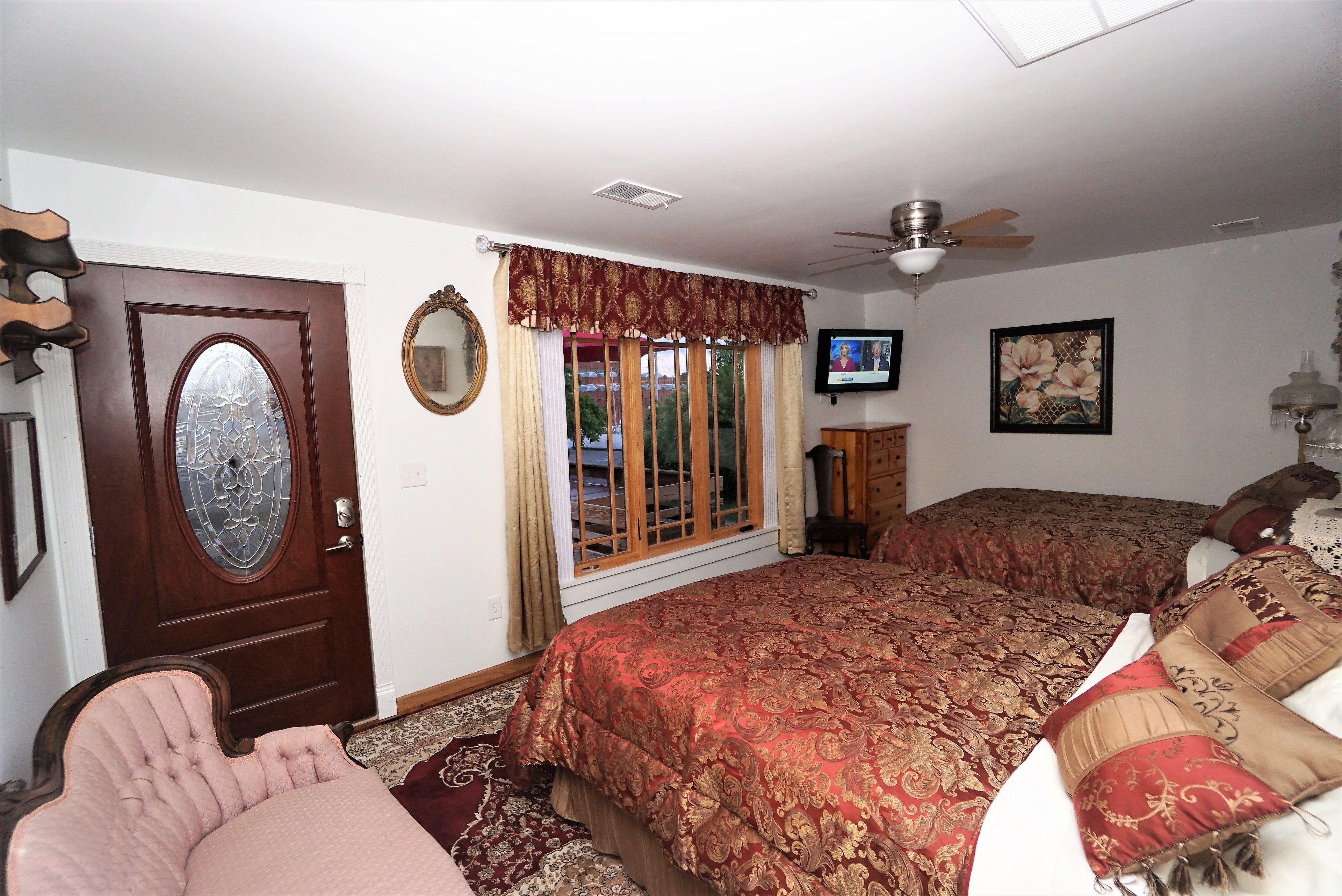 A bedroom with a large bed, a ceiling fan, a TV, and a window with curtains.