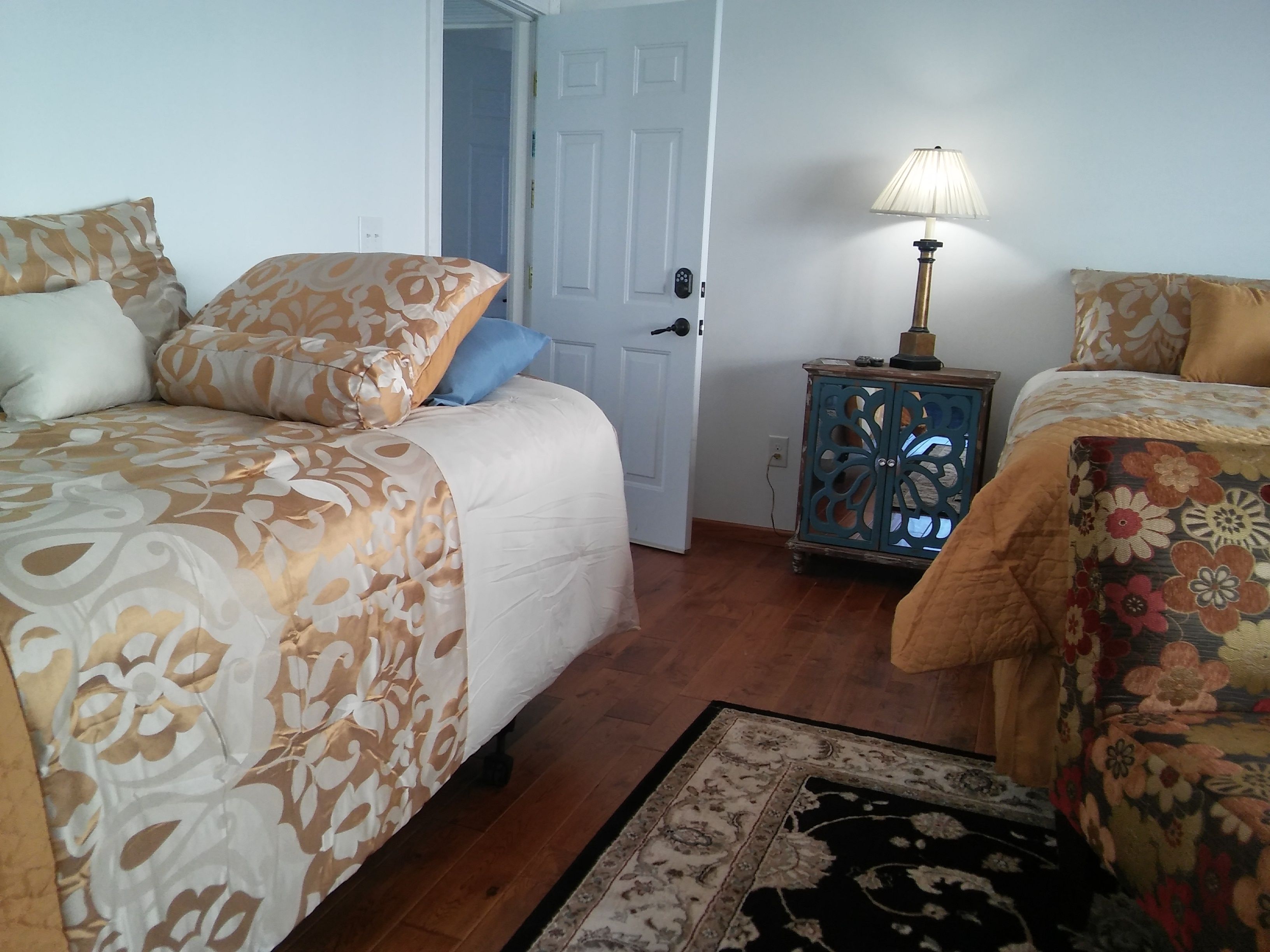 A bedroom with two beds, a nightstand with a lamp, and a door leading to another room.