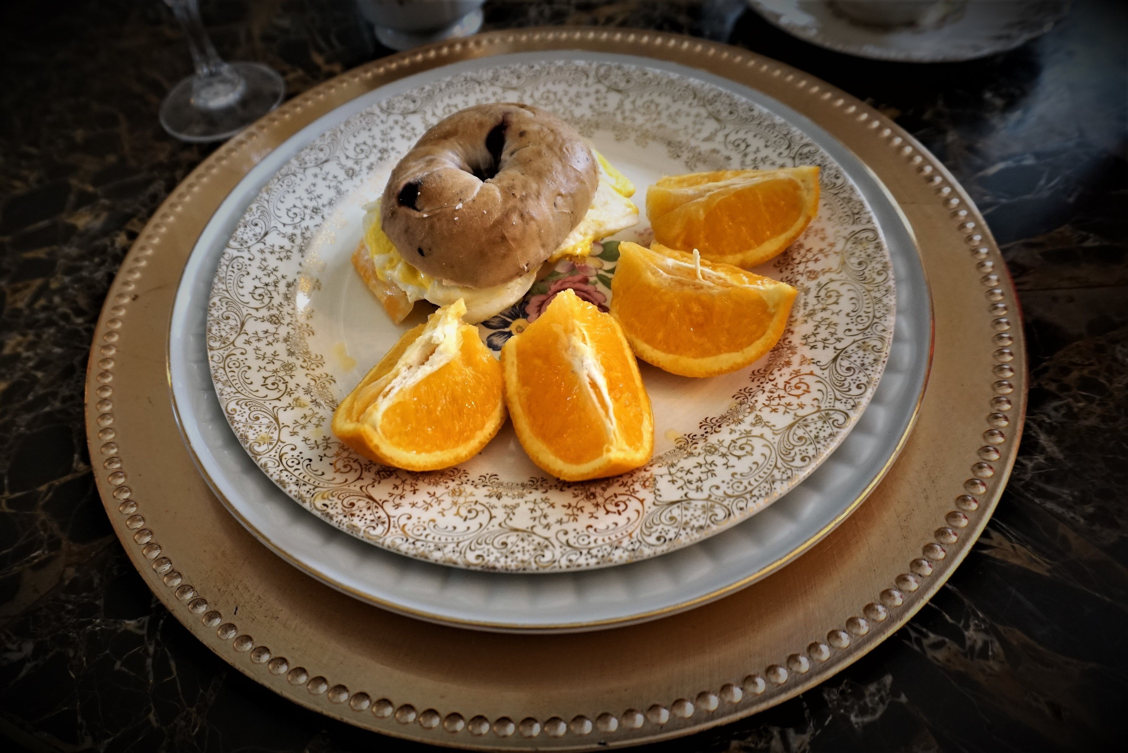 Une assiette contenant un bagel, des tranches d’orange et une boisson, posée sur une table en marbre.