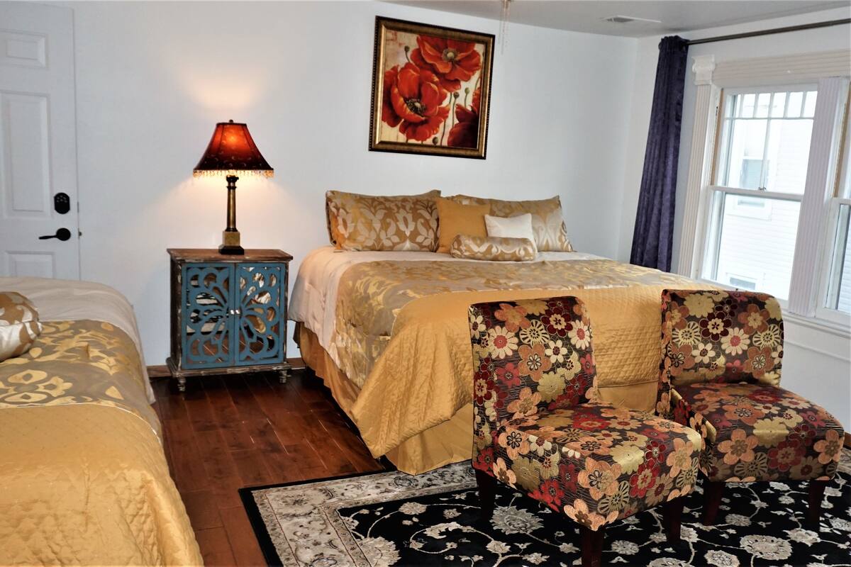 Une chambre à coucher avec un lit, une table de chevet avec une lampe, un fauteuil à motifs floraux et une grande fenêtre avec des rideaux.