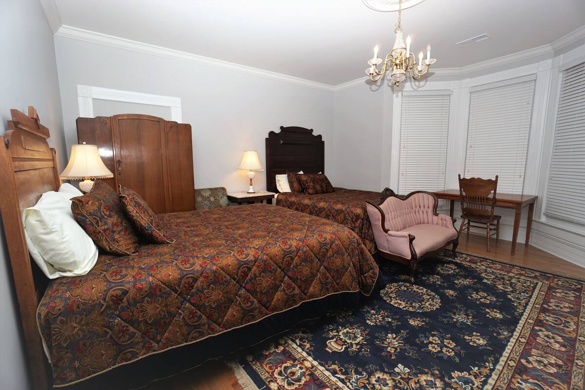 Une chambre avec deux lits, un lustre, une armoire en bois, une table avec des chaises et un tapis à motifs.