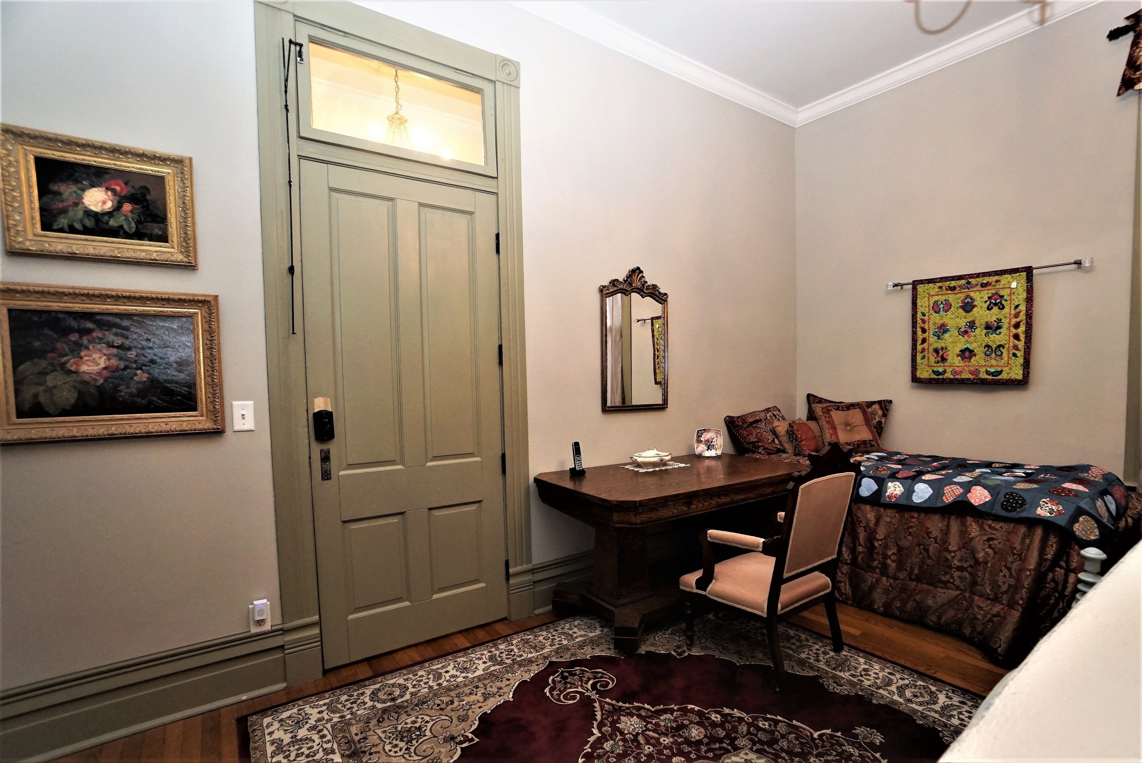 Une pièce comprenant un bureau en bois, une chaise, un lit avec une couverture, un miroir et deux tableaux encadrés accrochés au mur.