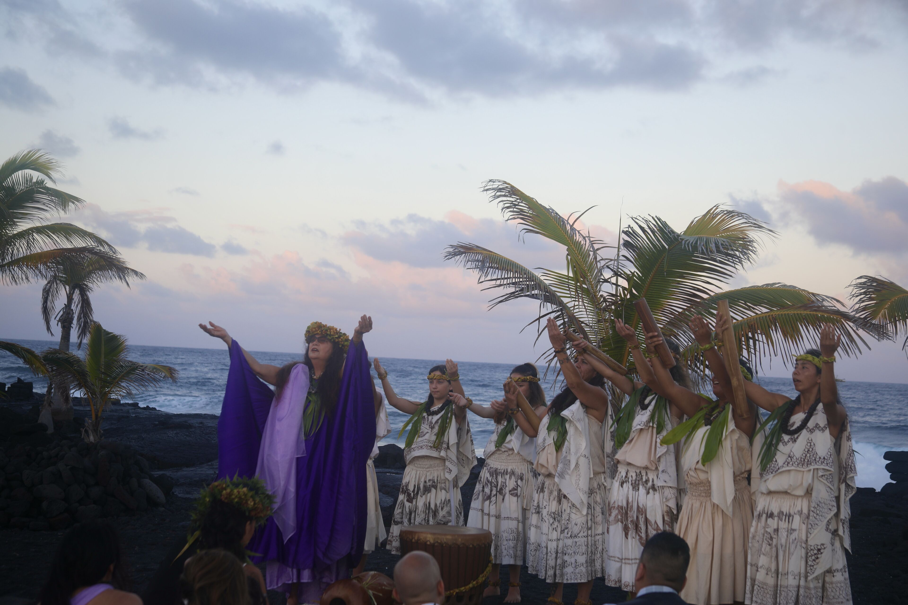 A group of people performing a traditional dance on a beach with palm trees and the ocean in the background.