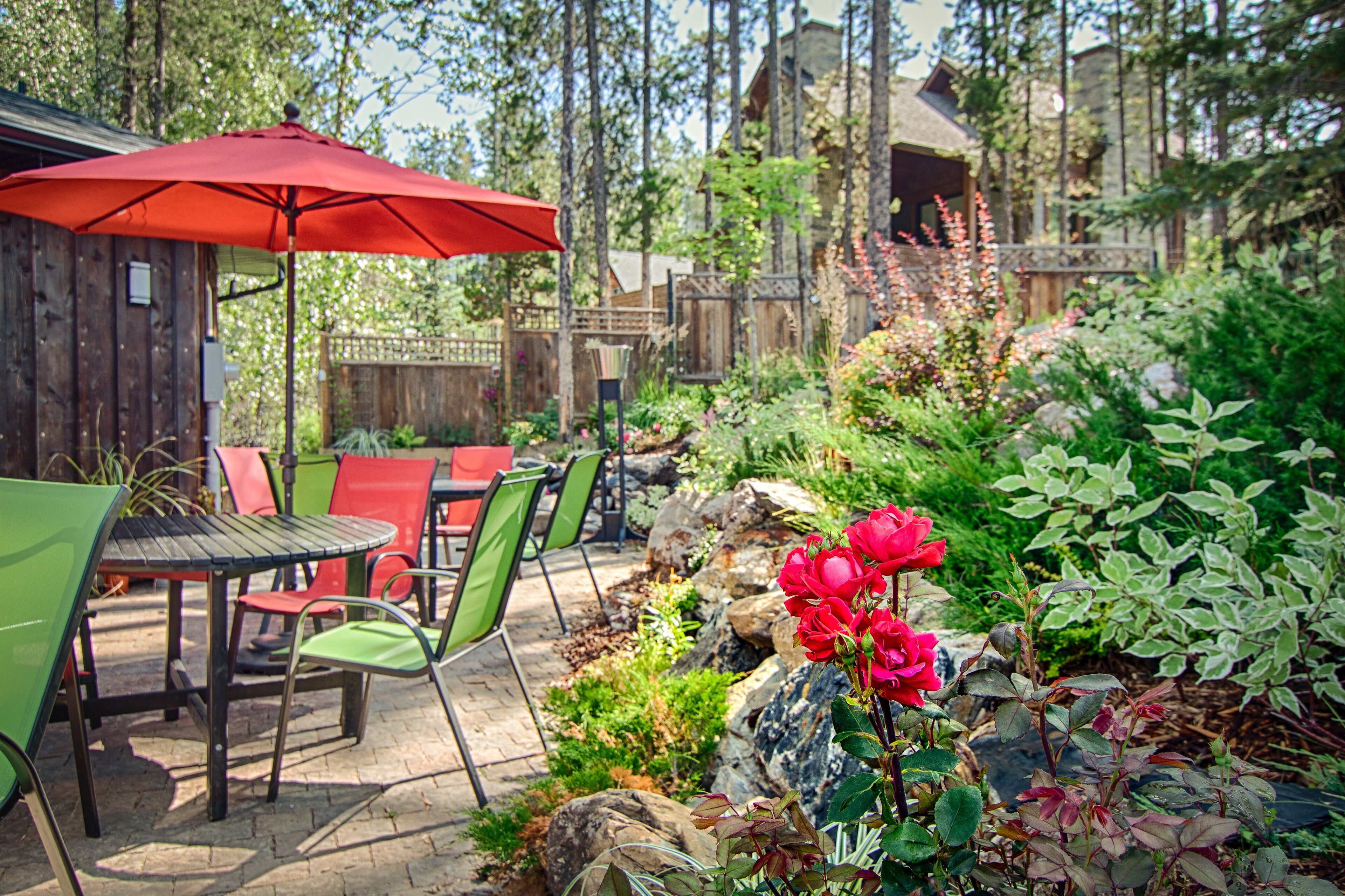 An outdoor patio with a red umbrella, green and red chairs, and a variety of plants and flowers.