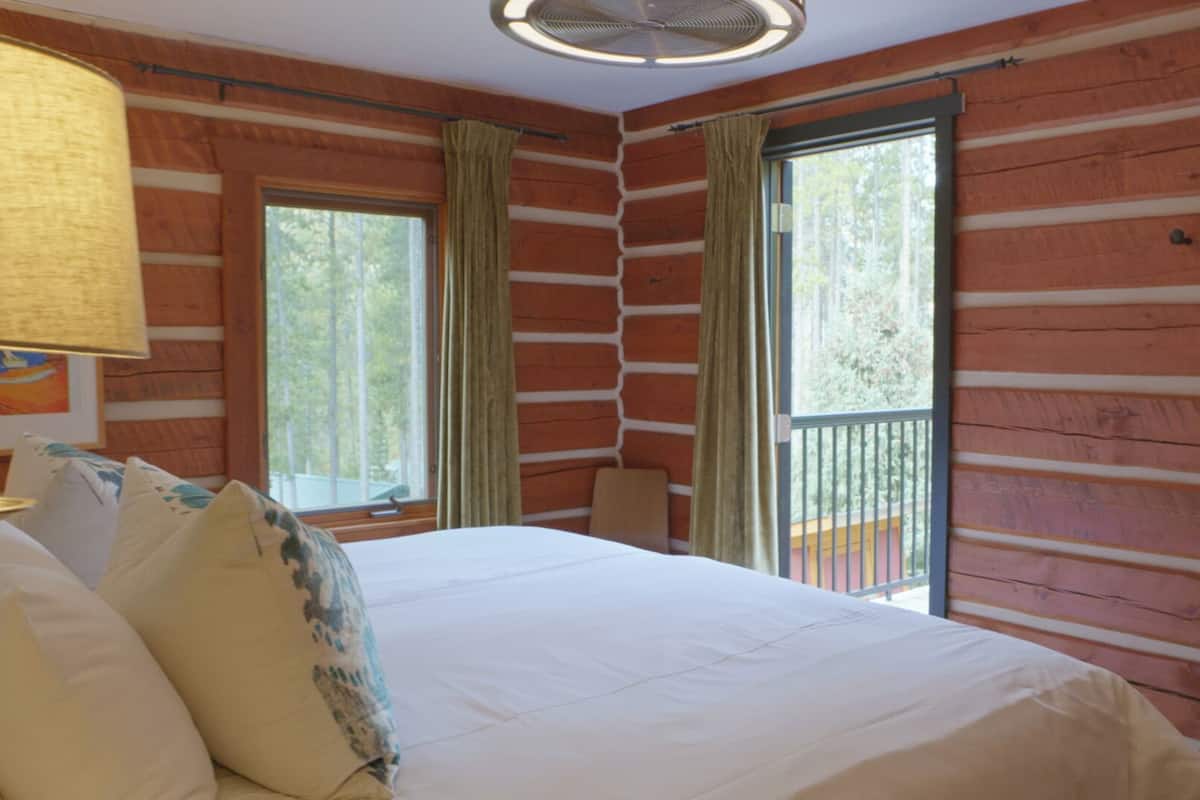 Habitación tipo cabaña de madera con una cama, dos ventanas, una silla y una lámpara.