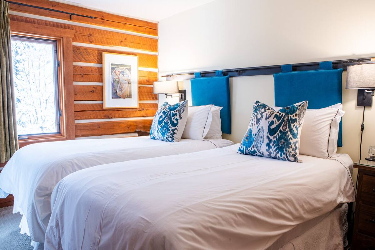 Habitación de hotel con dos camas, paneles de madera en las paredes, un cuadro enmarcado y una ventana con cortinas.
