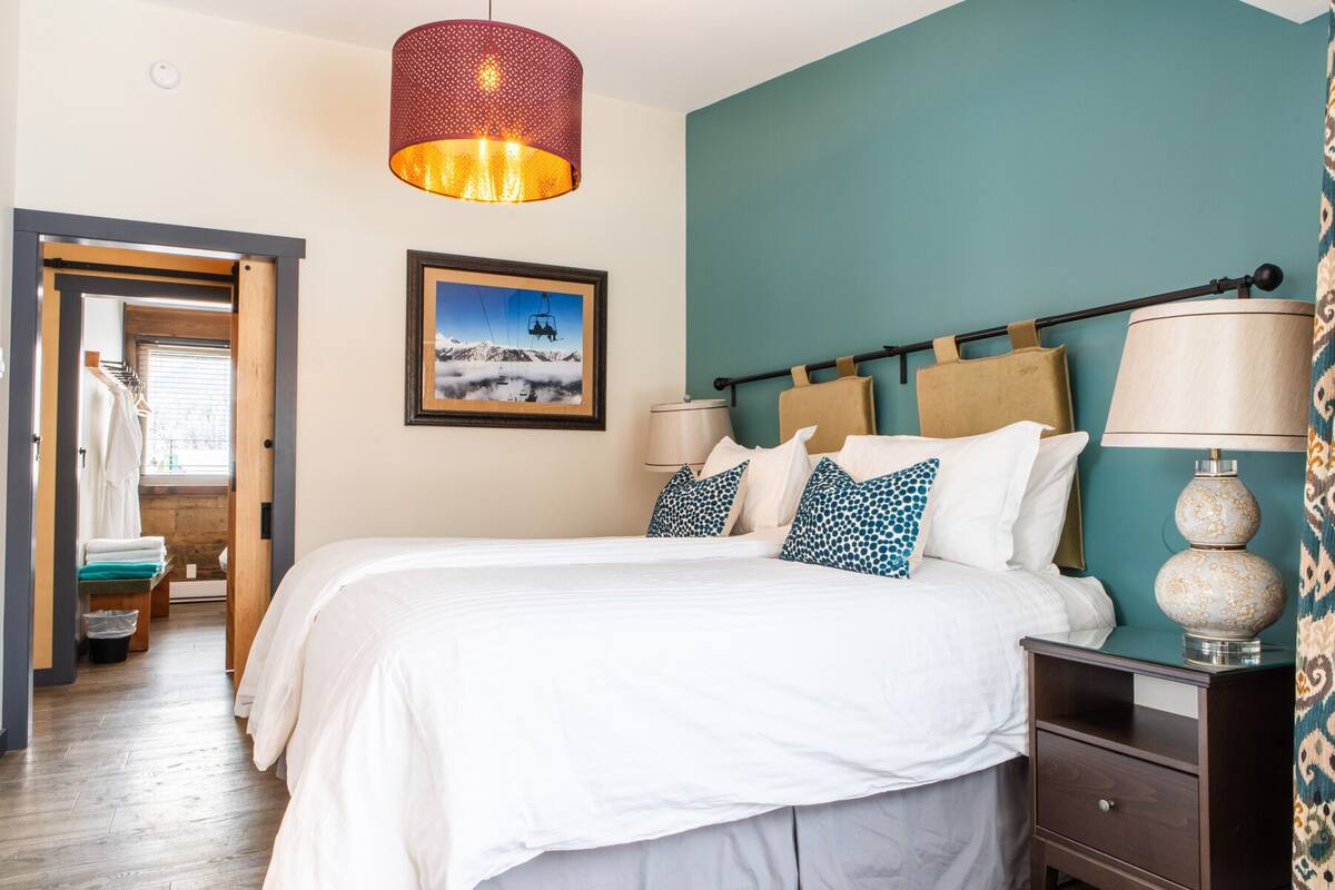 Una habitación de hotel con una cama, una mesita de noche, una lámpara, un cuadro de una montaña y una puerta que da a un baño.