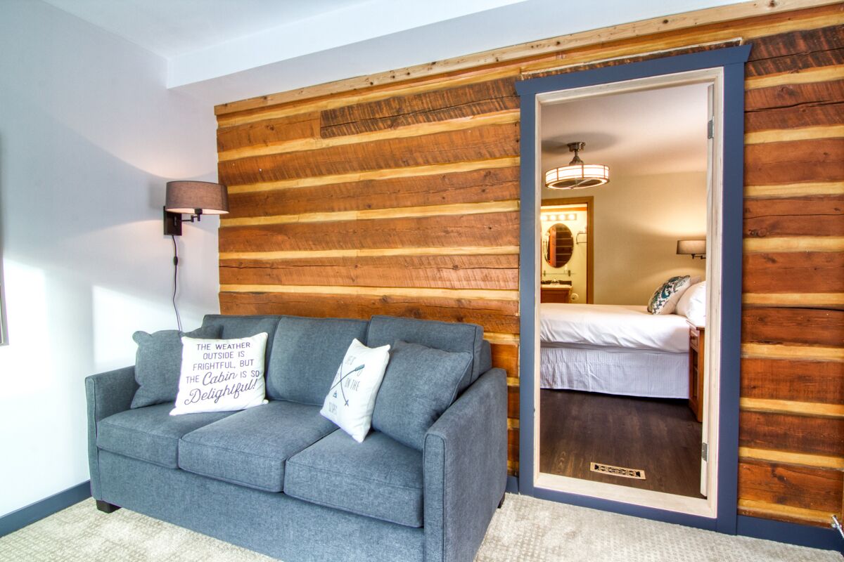 Interior acogedor de una cabaña con sofá gris, pared de madera, cama y espejo.