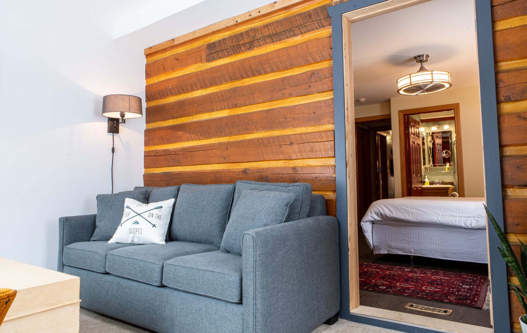 A room with a grey sofa, a wooden slat wall, a bed with a red rug, and a ceiling fan.