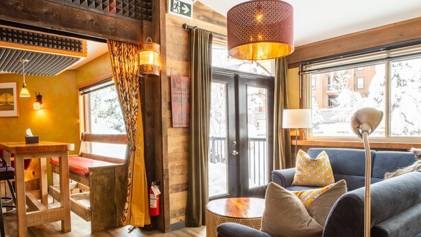 A cozy room with a wooden ceiling, a blue sofa with patterned cushions, a round wooden table, and a large window with a view of buildings outside. - Earl Grey Lodge (Panorama)