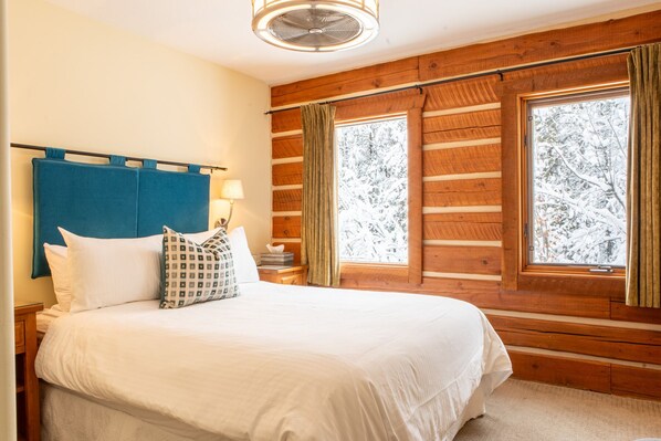 A bedroom with a wooden log wall, a bed with white bedding, a blue headboard, and two windows with curtains.