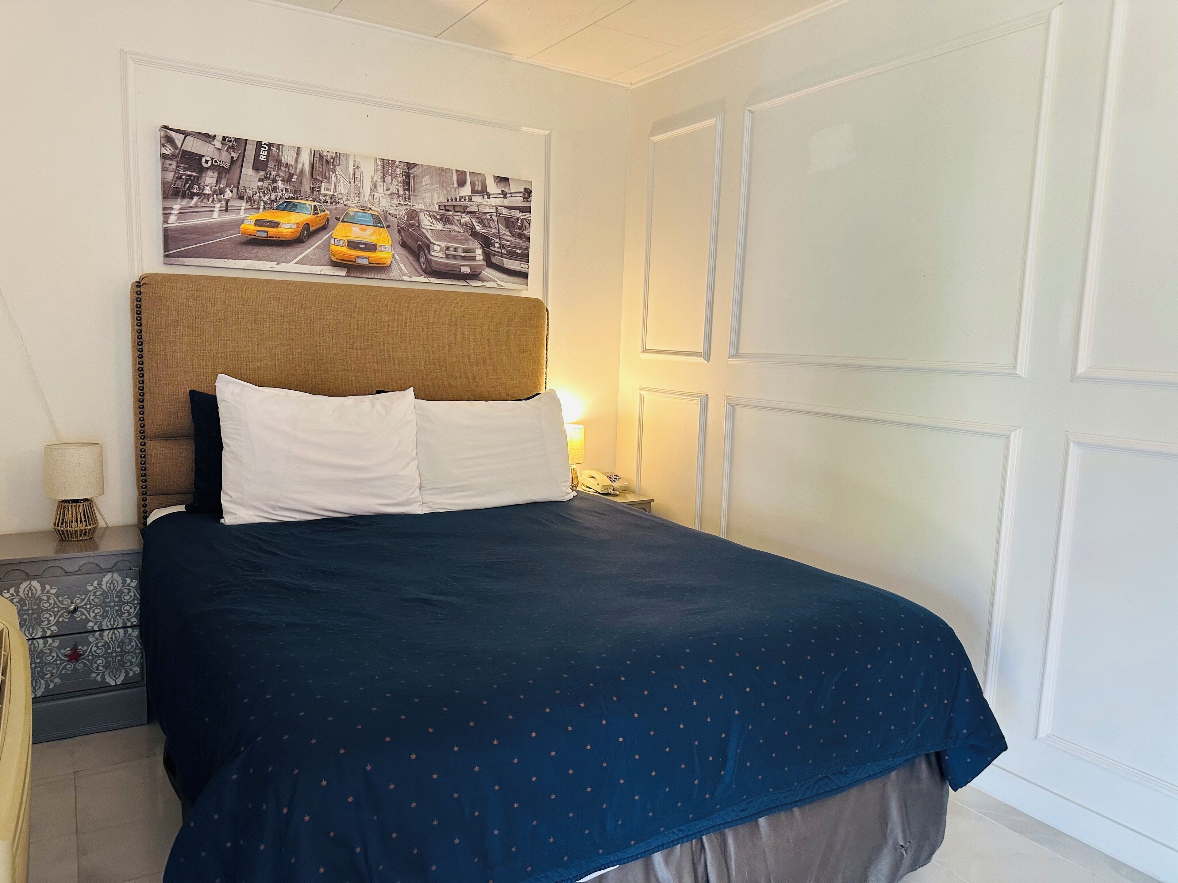 A hotel room with a bed, a headboard with a cityscape painting, a nightstand with a lamp, and a door.