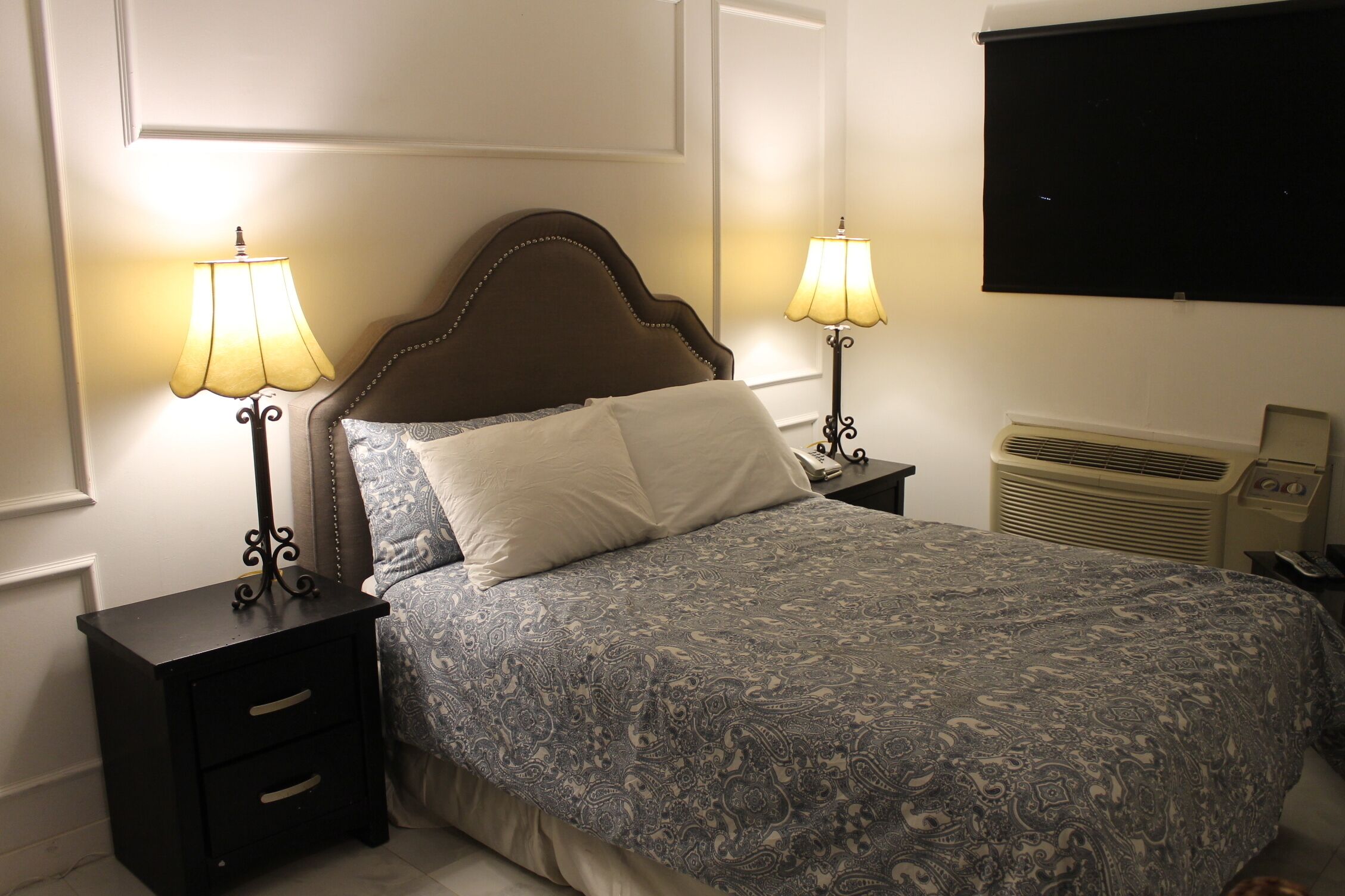 A neatly made bed with a patterned bedspread, two bedside tables with lamps, a wall-mounted air conditioner, and a plain wall.