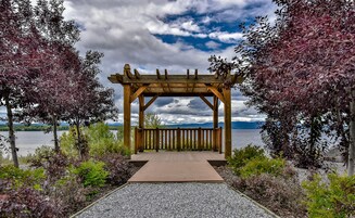 Uma pérgola de madeira com um caminho de cascalho que a conduz, cercada por árvores e um corpo d'água ao fundo.