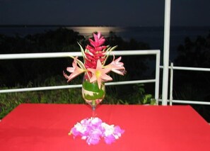 Une table recouverte d’une nappe rouge, un vase en verre contenant des fleurs roses et un verre d’eau.