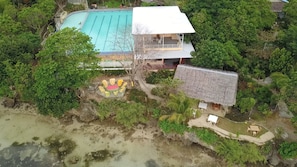 Vue aérienne d’un complexe hôtelier avec une piscine, une structure à toit de chaume et une végétation luxuriante.