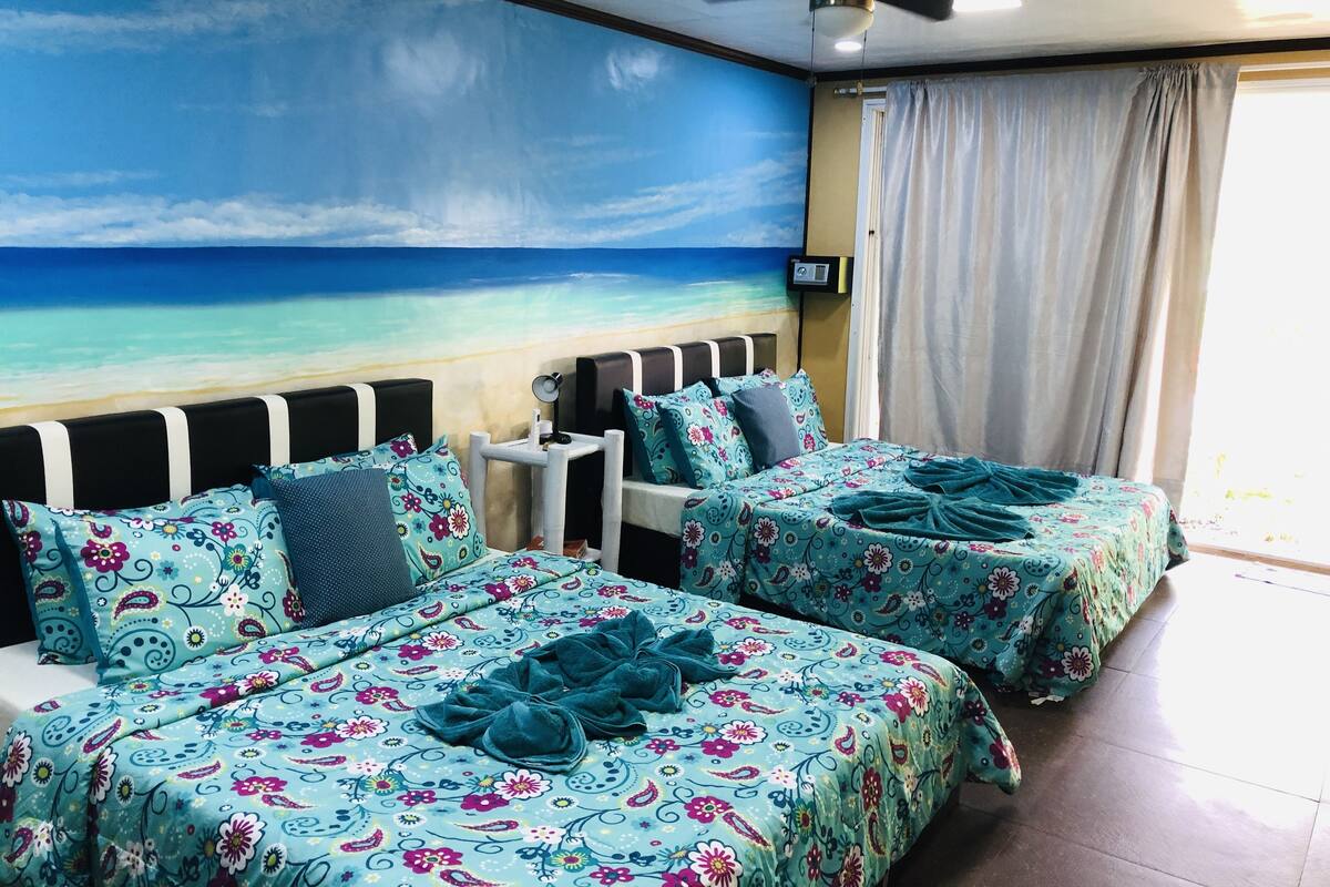 Une chambre avec deux lits, un ventilateur de plafond, une fresque murale sur le thème de la plage et une fenêtre avec des rideaux.