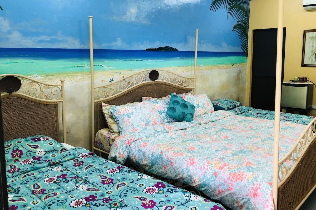 Une chambre dotée d’un lit orné d’une fresque représentant une plage tropicale, d’une tête de lit en osier et d’une literie à motifs floraux.