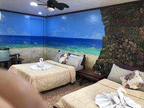 Une chambre d’hôtel avec deux lits, un ventilateur de plafond, une table de chevet et une fresque murale représentant une scène de plage.