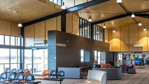 A spacious airport departure lounge with seating areas, a check-in counter, and large windows providing views of the outside.