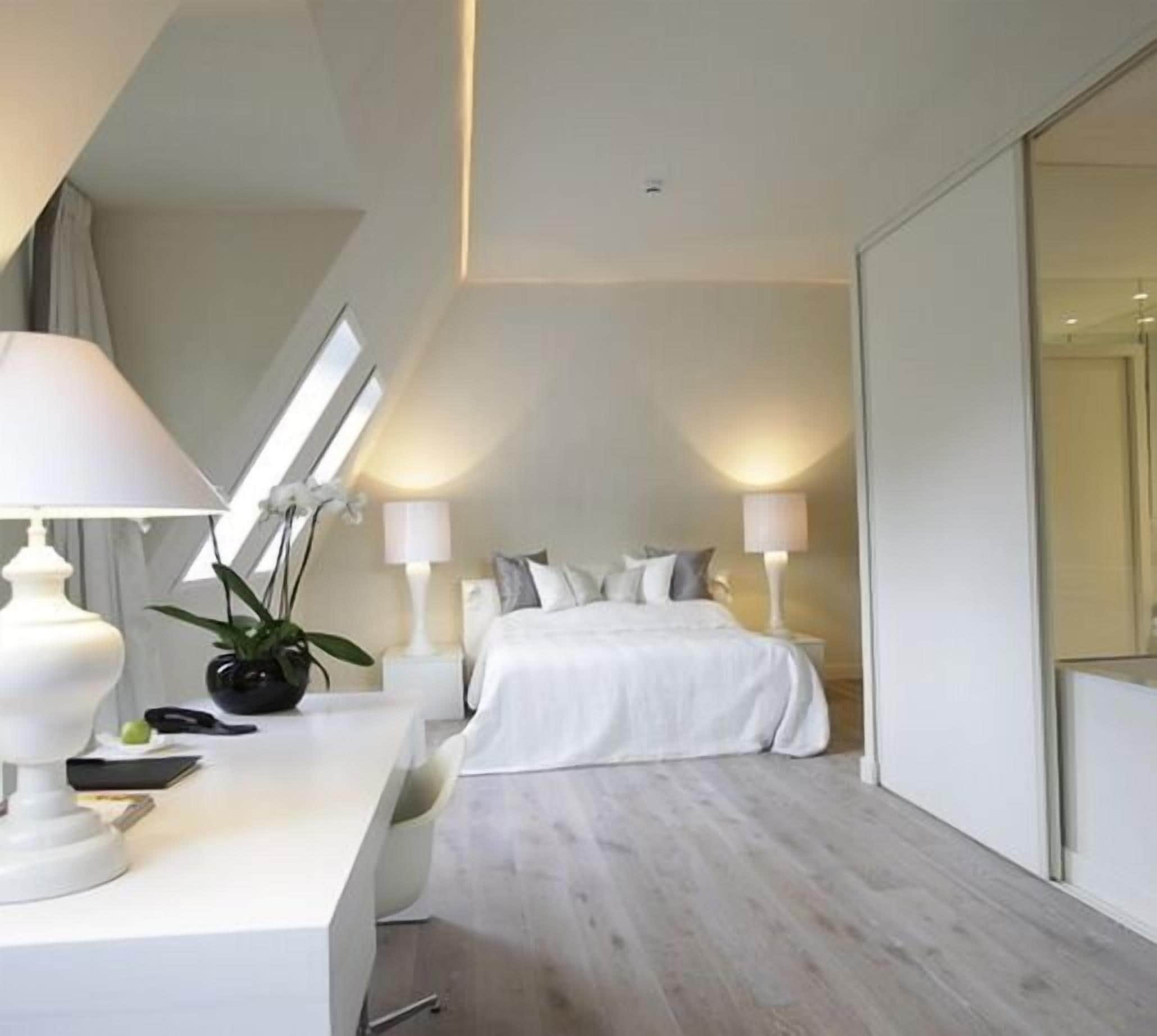 A modern hotel room with a white bed, a desk with a lamp, and a large window providing natural light.