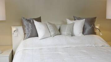 Una cama bien hecha con ropa de cama blanca y varios almohadones, una mesita de noche con una lámpara, y una lámpara de pared.