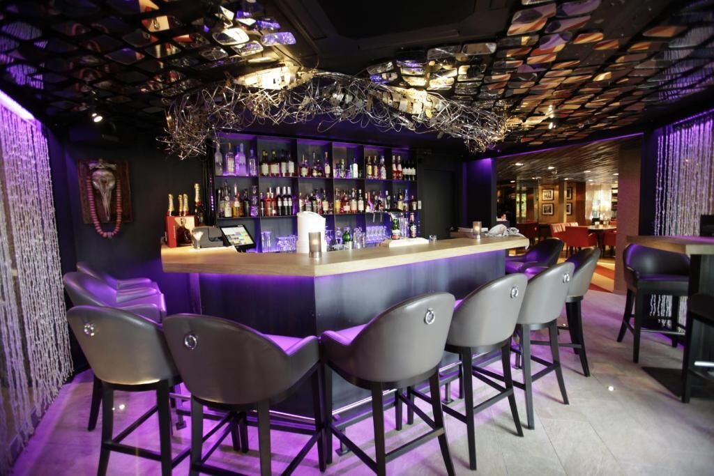 Zona de bar con mostrador, varios taburetes y estantes llenos de botellas.