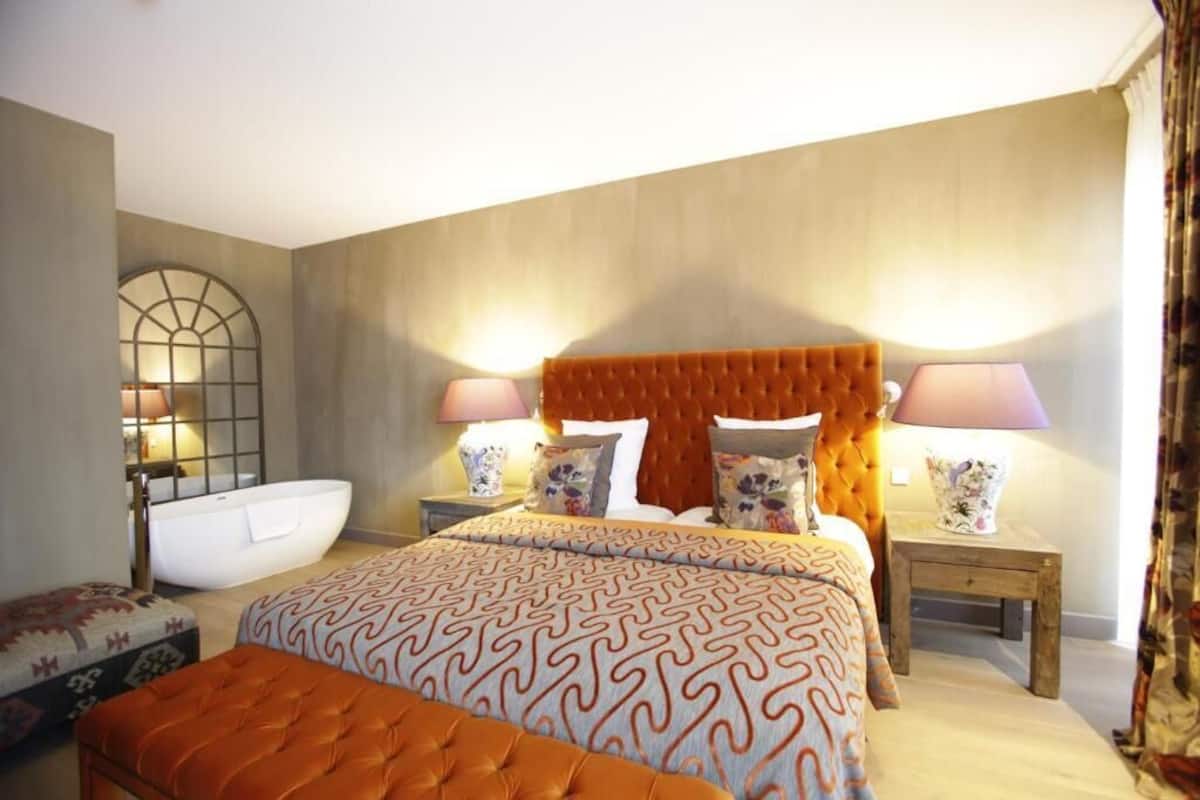 A bedroom with a large bed, two bedside tables with lamps, a white bathtub, and a wooden bench.