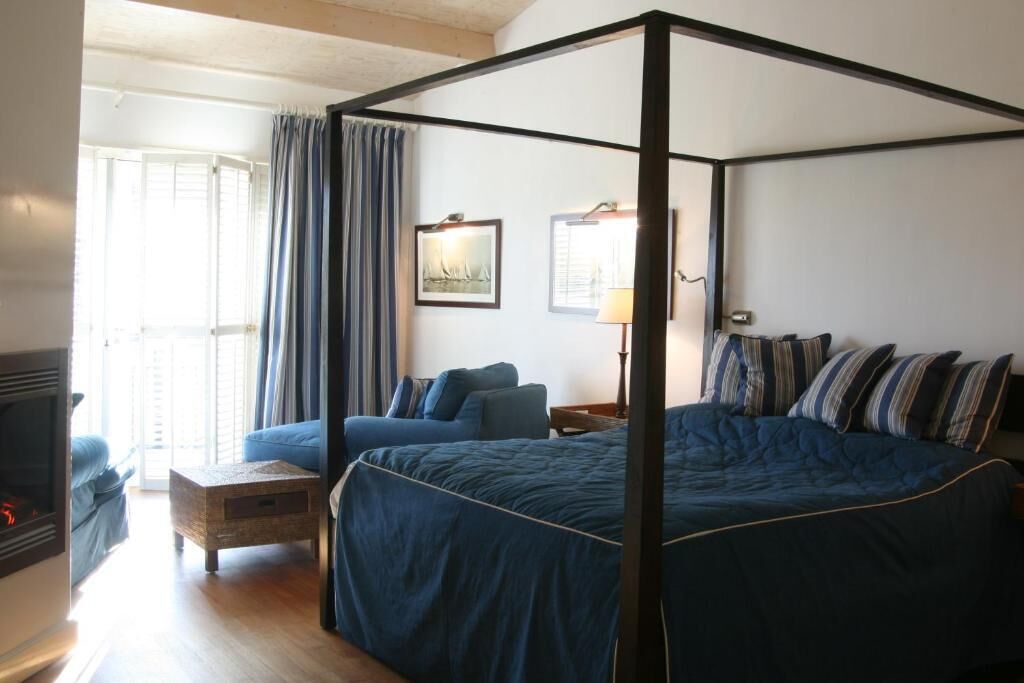 A four-poster bed with a dark blue bedspread and striped pillows, a wooden nightstand with a lamp, a blue sofa, and a fireplace.