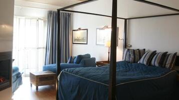 A four-poster bed with a dark blue bedspread and striped pillows, a wooden nightstand with a lamp, a blue sofa, and a fireplace.